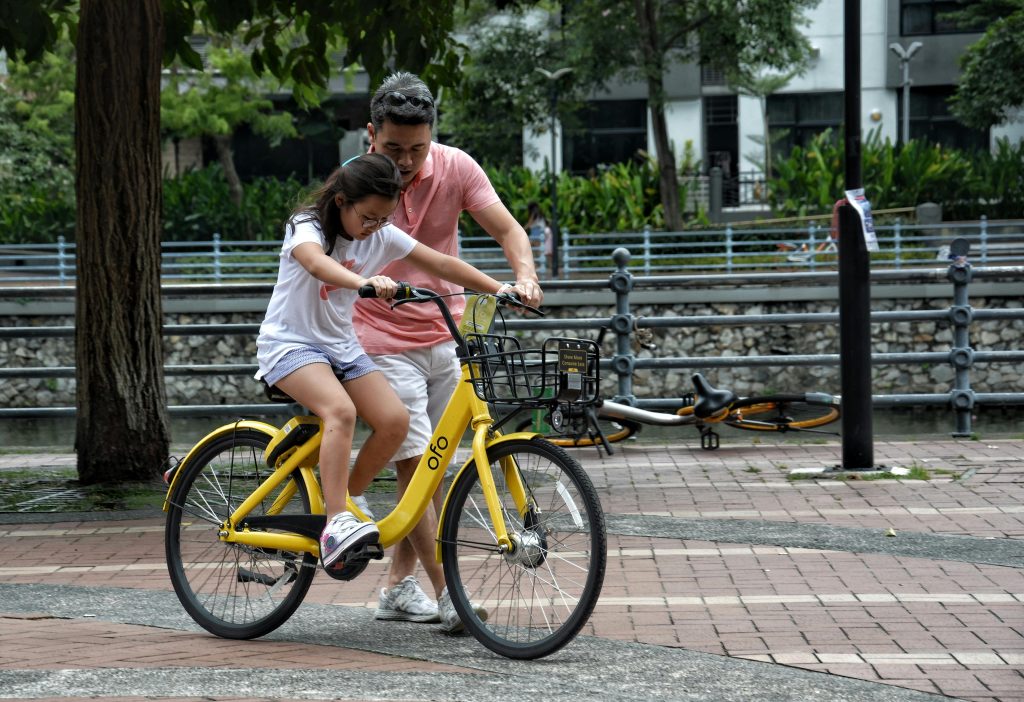 Father teaches daughter to ride a yellow bicycle in a Singapore park. Bonding moment.